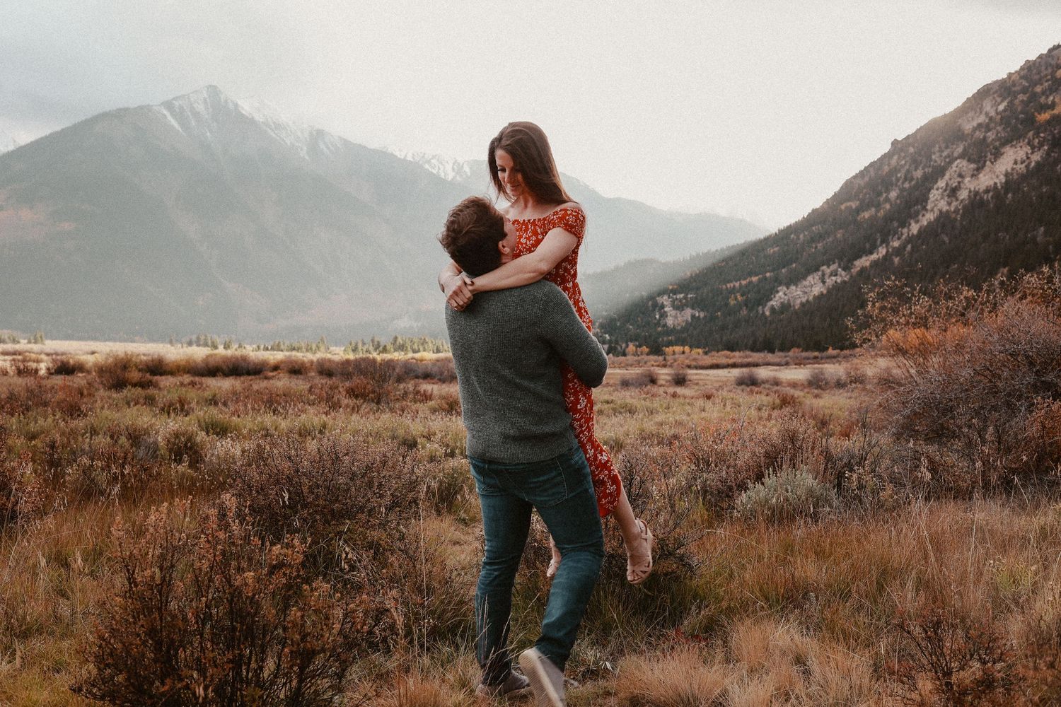 Two people embrace in a romantic moment against a backdrop of majestic mountains and autumn brush in a scenic valley.