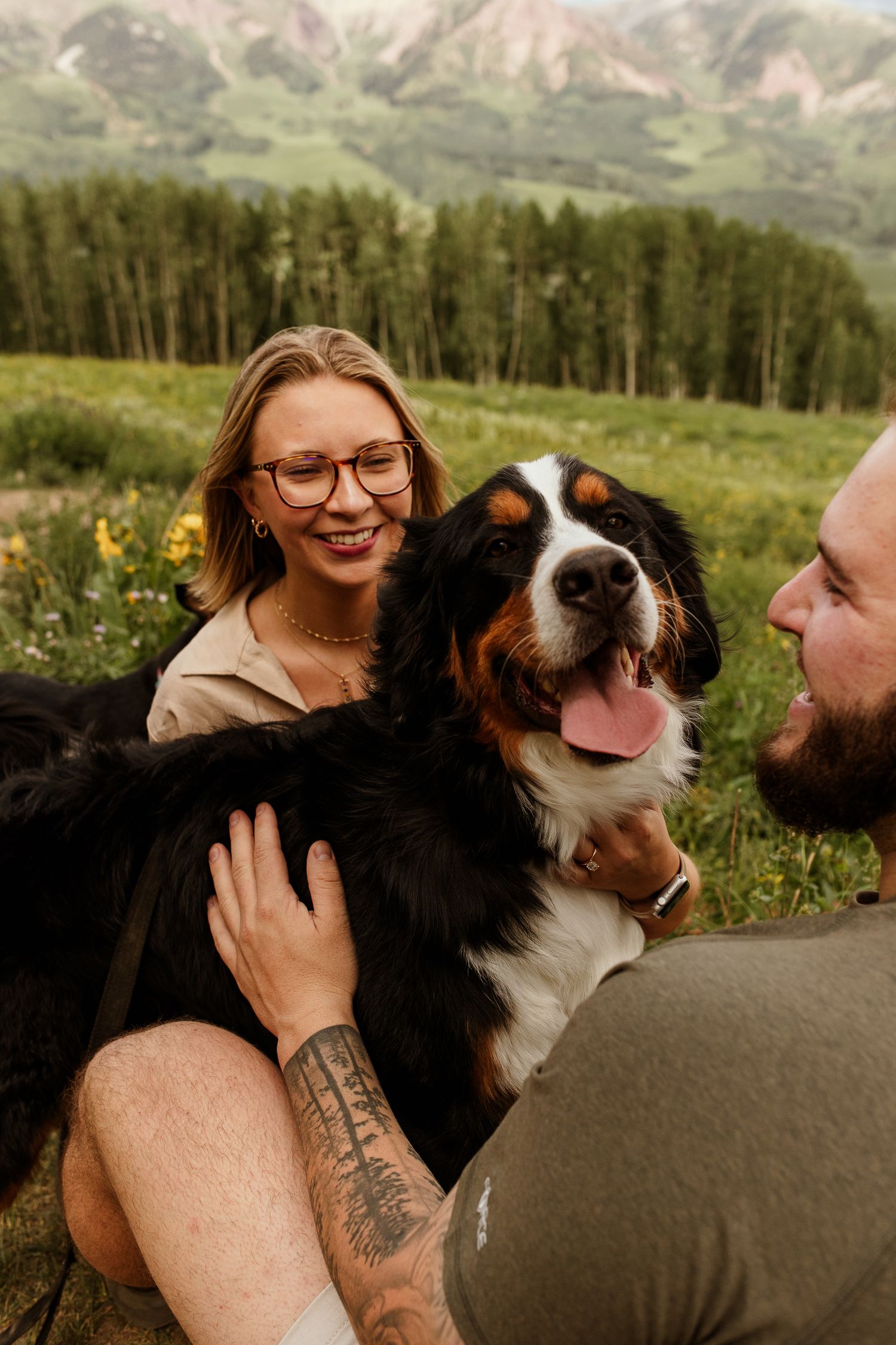 A couple shares joyful moments with their black and white Australian Shepherd dog against a mountain backdrop.
