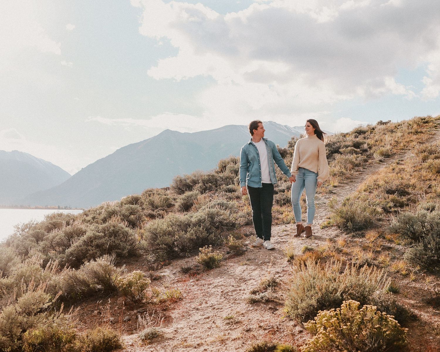 A couple hiking along a desert mountain trail with scenic mountain views and sage brush in soft golden light.
