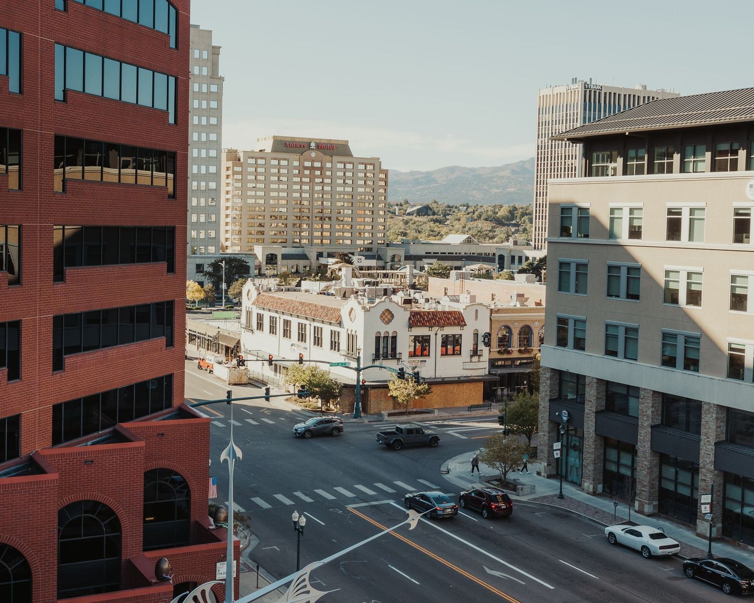Aerial view of a downtown city intersection with brick buildings, mountain backdrop, and light traffic on a sunny day.