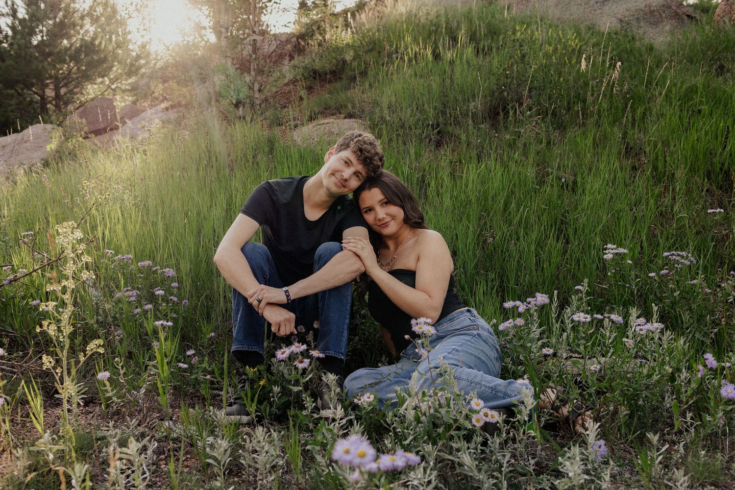 A couple sits together in a wildflower meadow at sunset surrounded by tall grass and blooming flowers.
