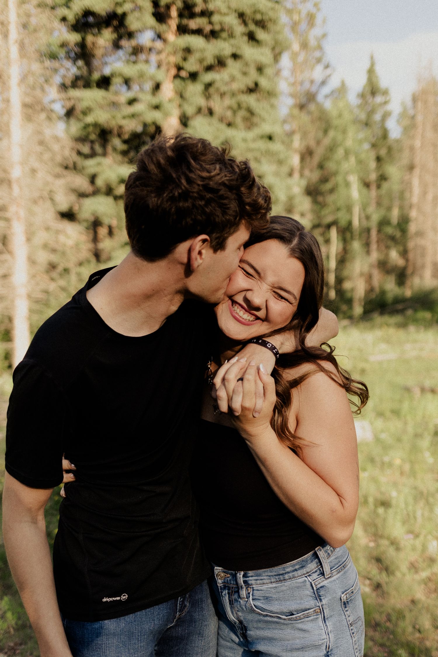 Couple in black tops embracing and laughing in a sunlit forest, sharing a joyful romantic moment outdoors.