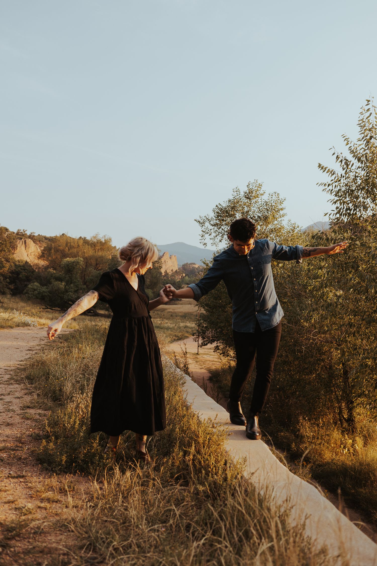 Two people balance while walking along a concrete barrier on a scenic hiking trail during golden hour.
