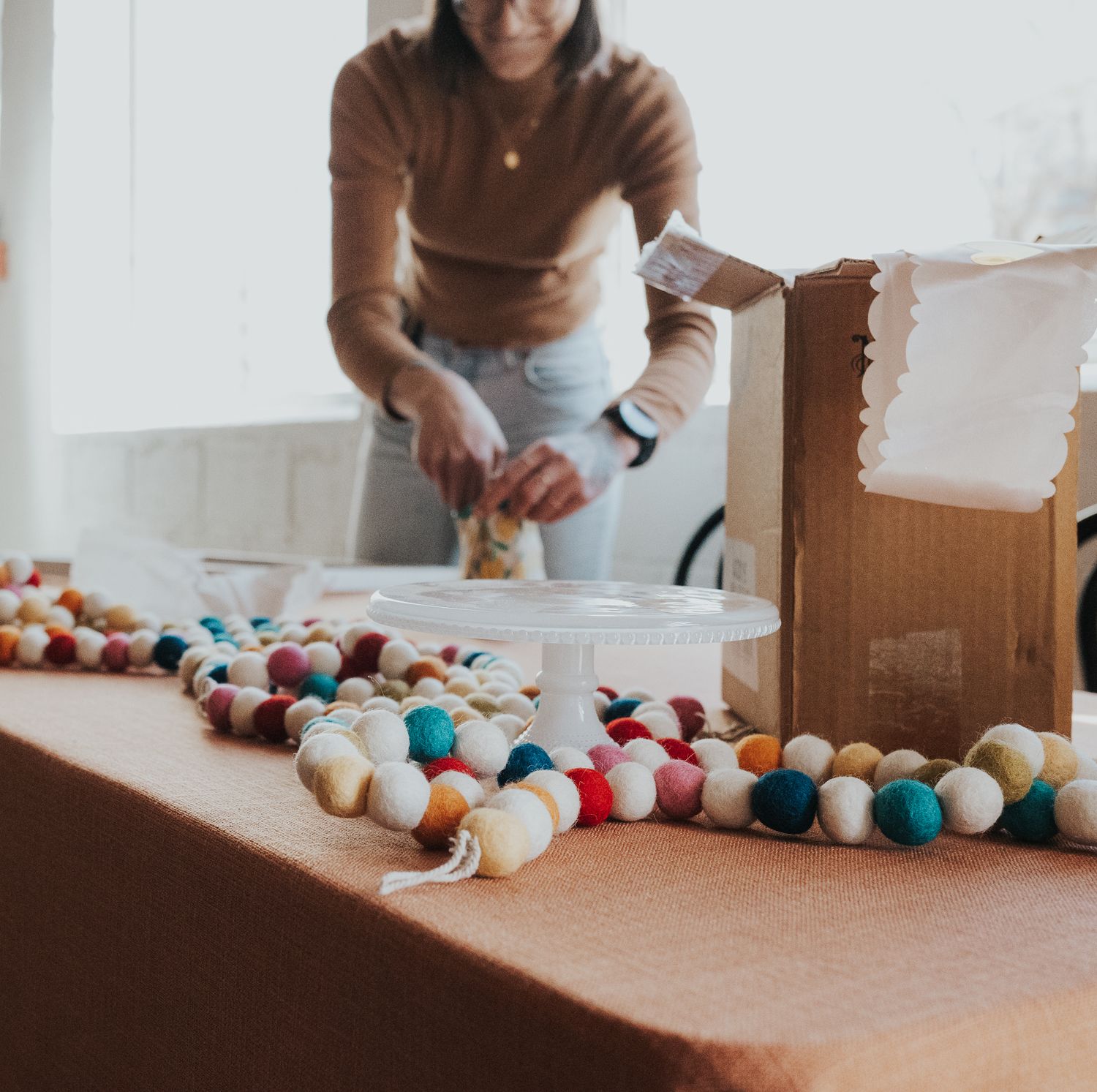 Colorful felt ball garland on a table with a woman unpacking a cardboard box in the background.