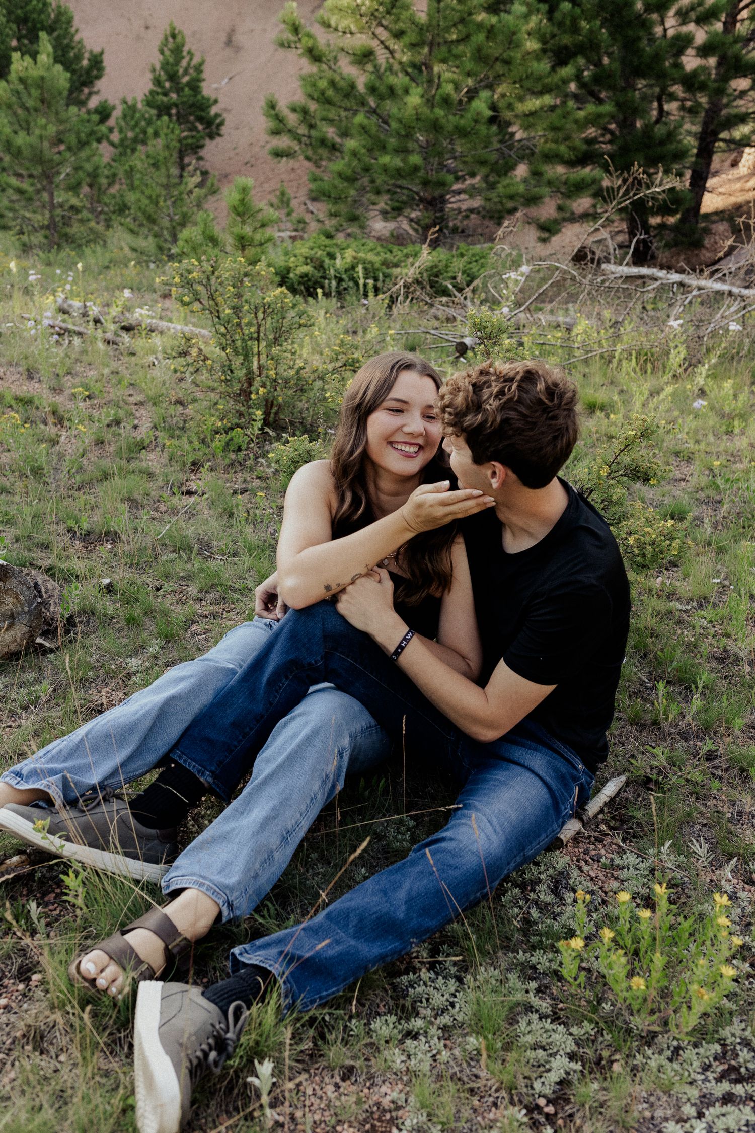 Couple sharing a joyful moment while sitting together on a grassy hillside with pine trees in the background.