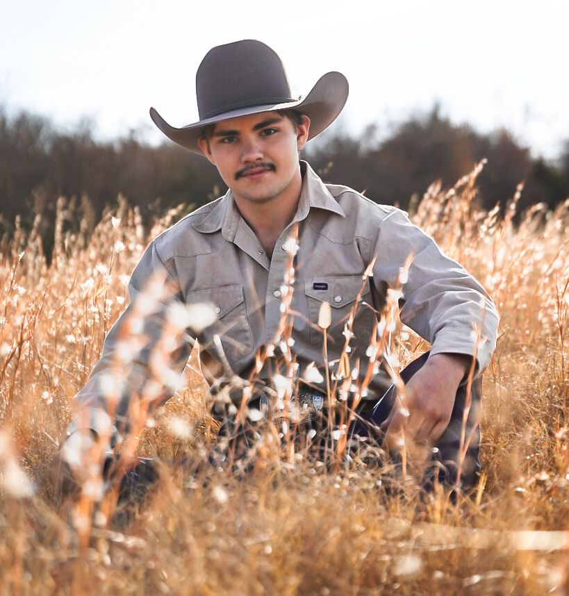Person in cowboy hat and western wear poses in tall golden grass.