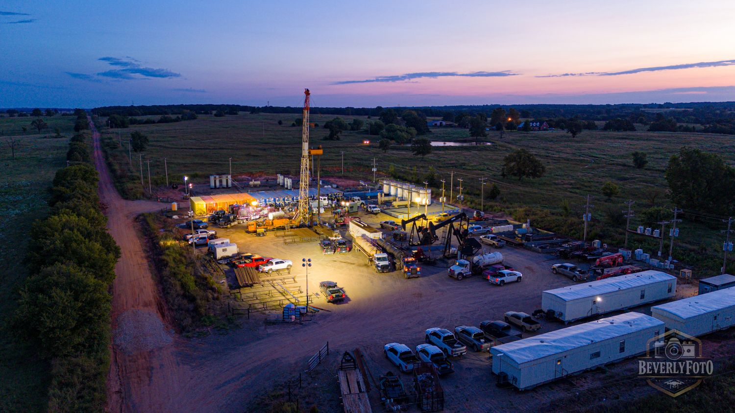 Aerial panoramic view of a small town's industrial area illuminated at dusk with glowing lights against darkening sky.