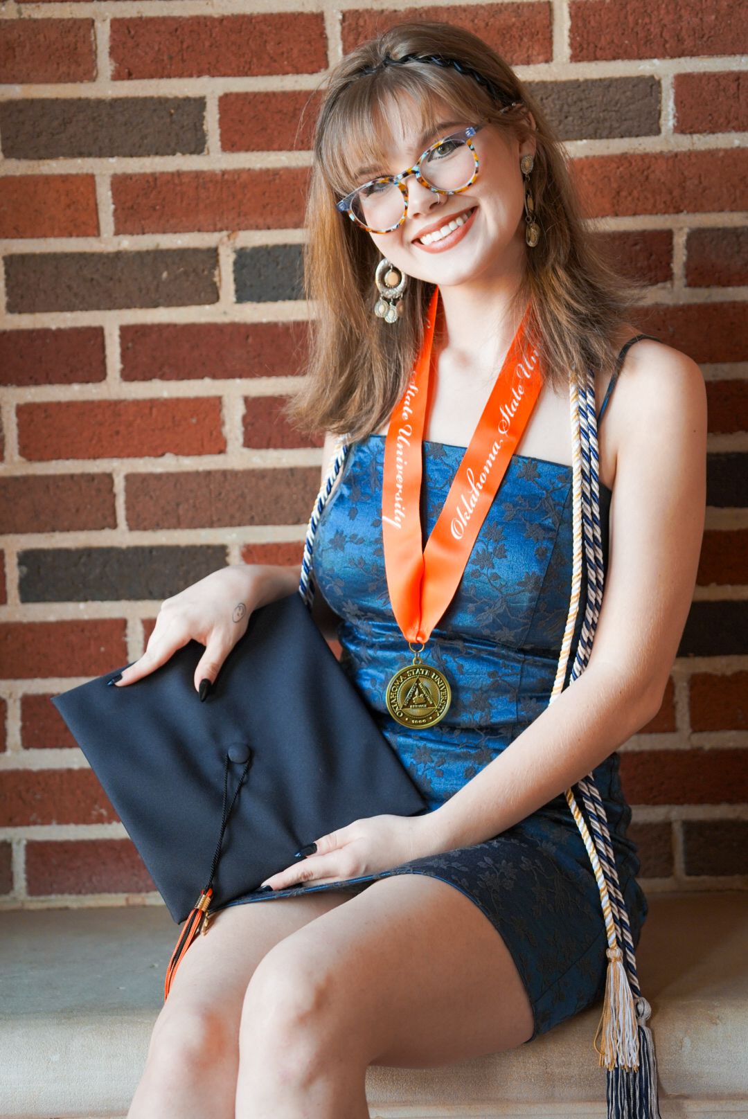 Graduate in blue dress displaying academic regalia against brick wall background.