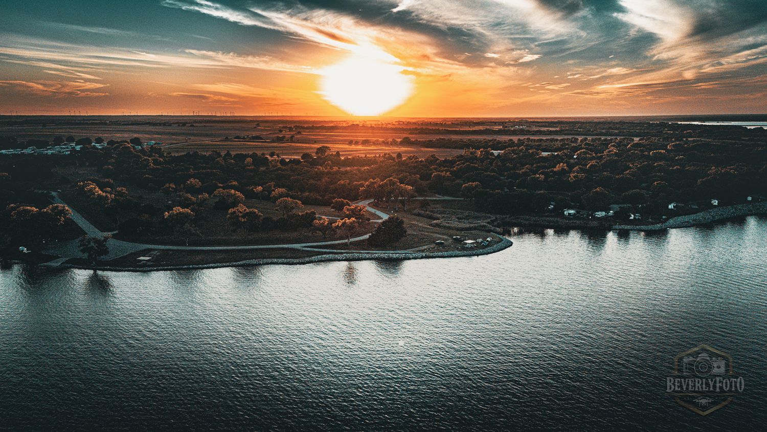 Stunning aerial panorama of a coastal sunset over calm waters with dramatic orange and teal clouds stretching across horizon.