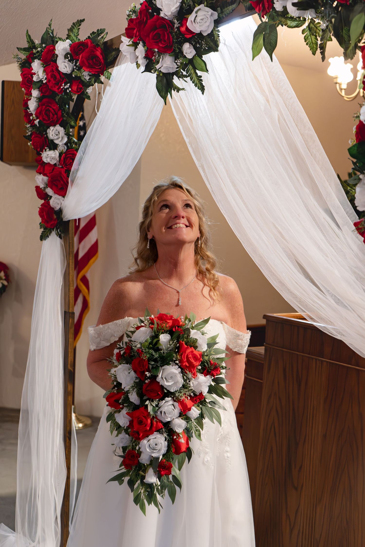 Wedding bouquet features vibrant red and white roses against flowing white drapery and floral arch decorations.