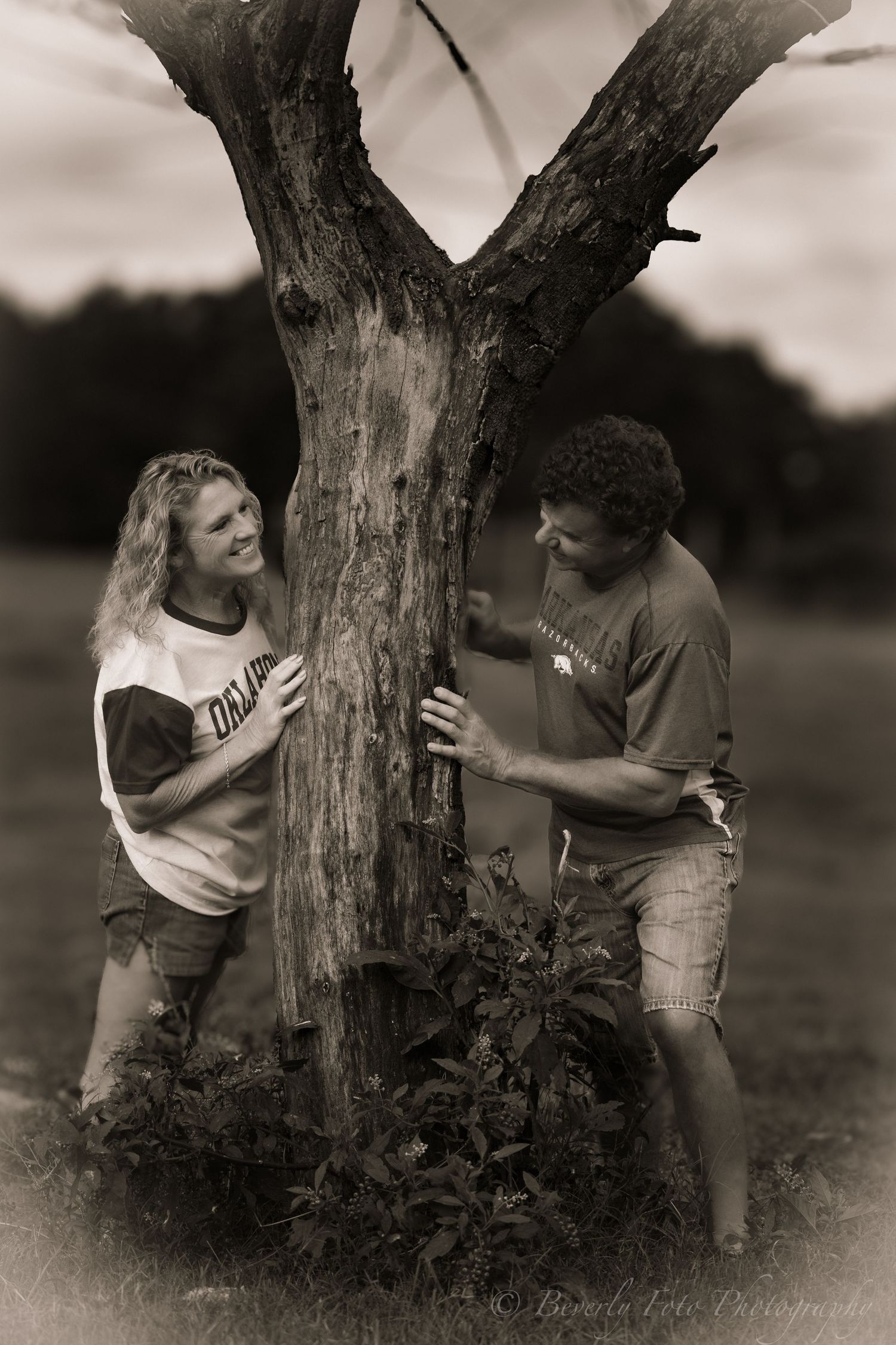 Artistic black and white photo of two people playfully interacting near a tree.
