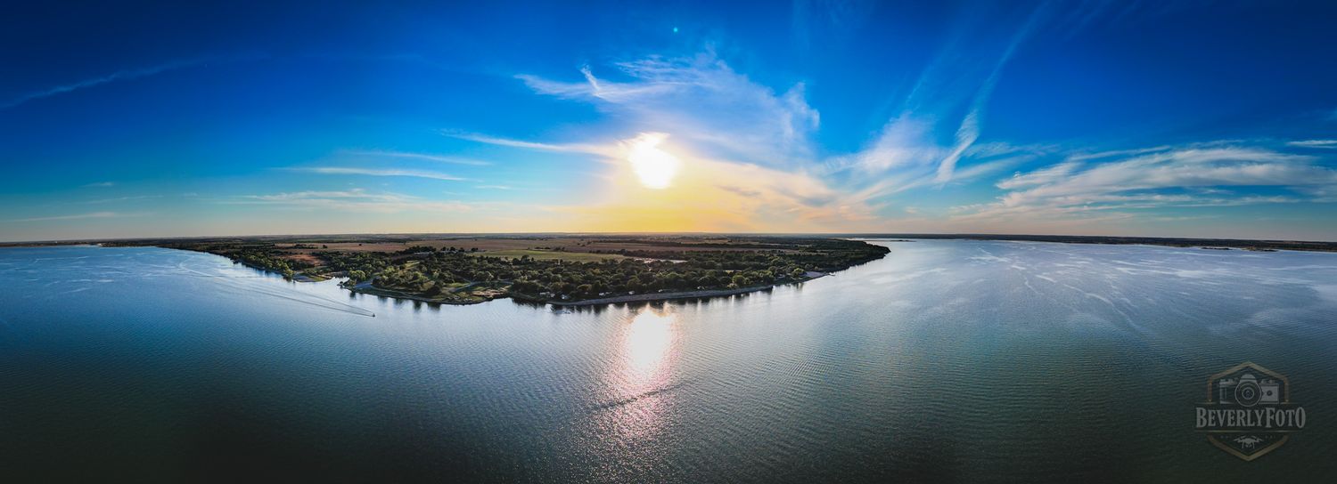 Panoramic aerial view of a serene lake surrounded by lush forests and mountains under a vibrant blue sky at sunset.
