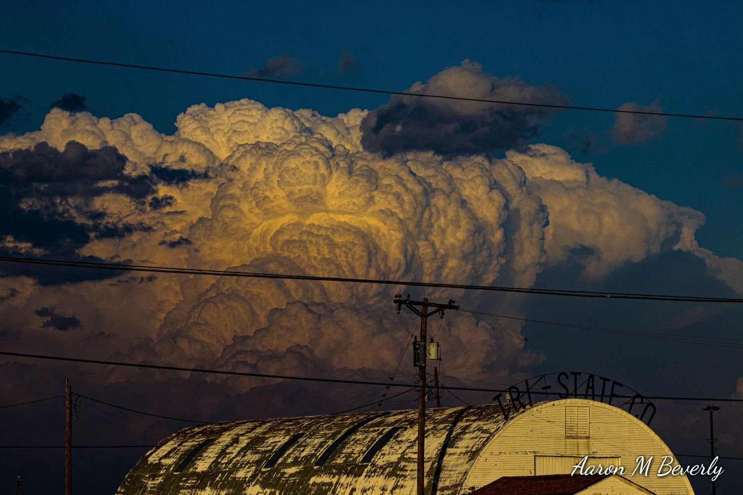 Dramatic golden clouds illuminate the sky above silhouetted power lines and buildings at sunset.