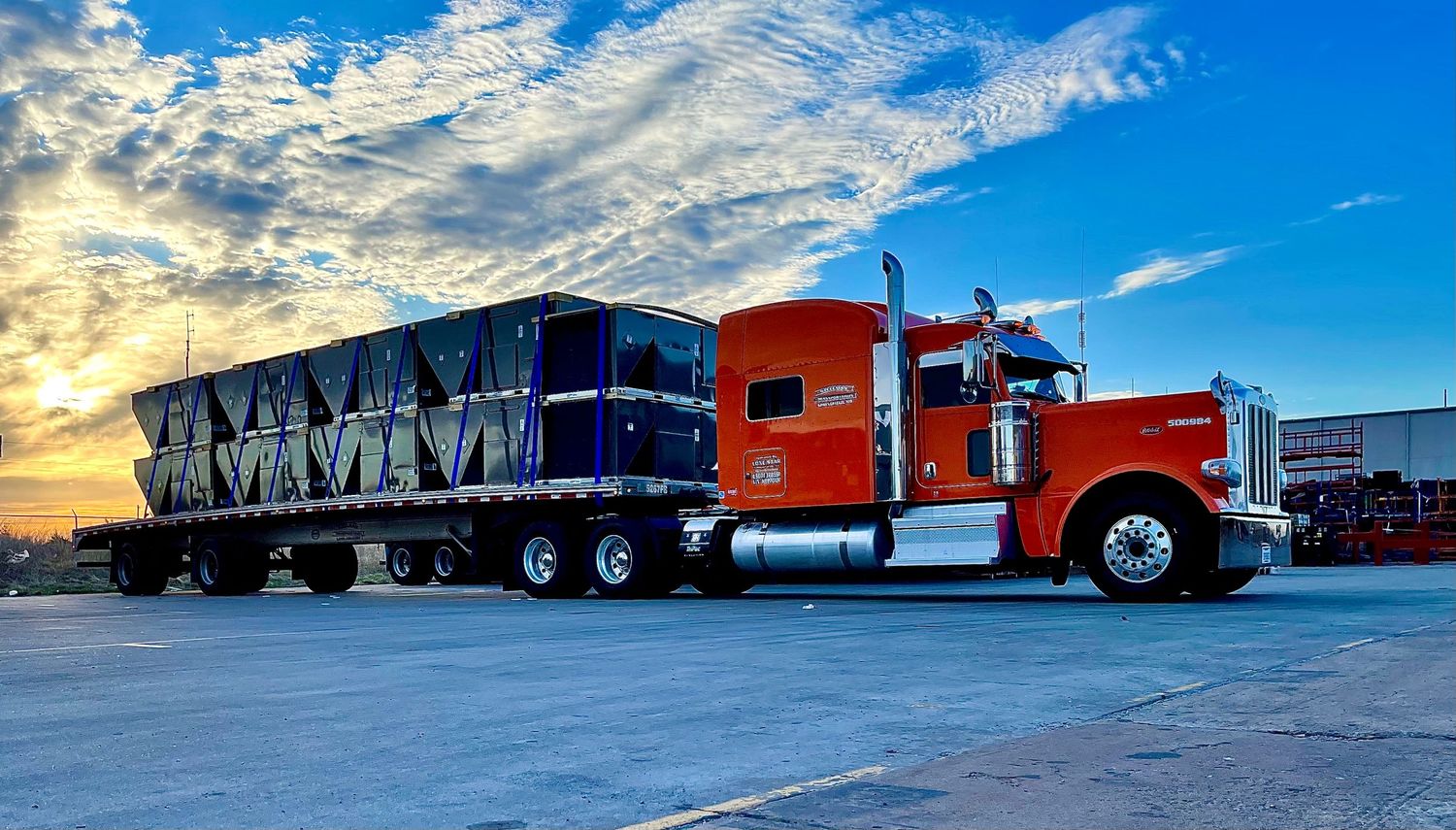 Red and blue semi truck with loaded trailer against vibrant sunset clouds.