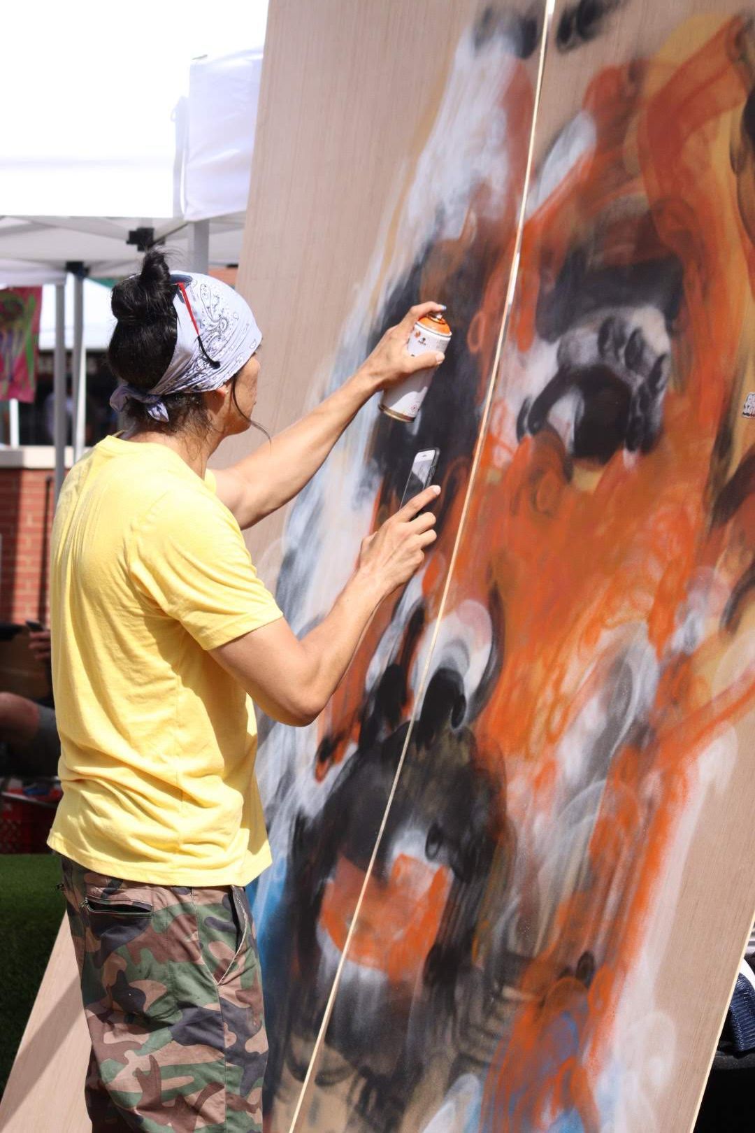 Artist in yellow shirt creating abstract painting with orange and black colors on large canvas.