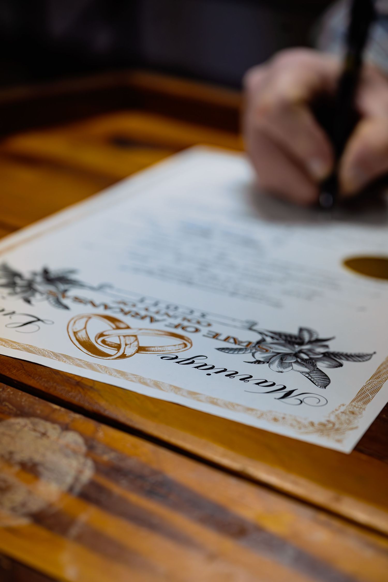 A hand writes on an elegant certificate with decorative floral designs and gold accents on a wooden surface.