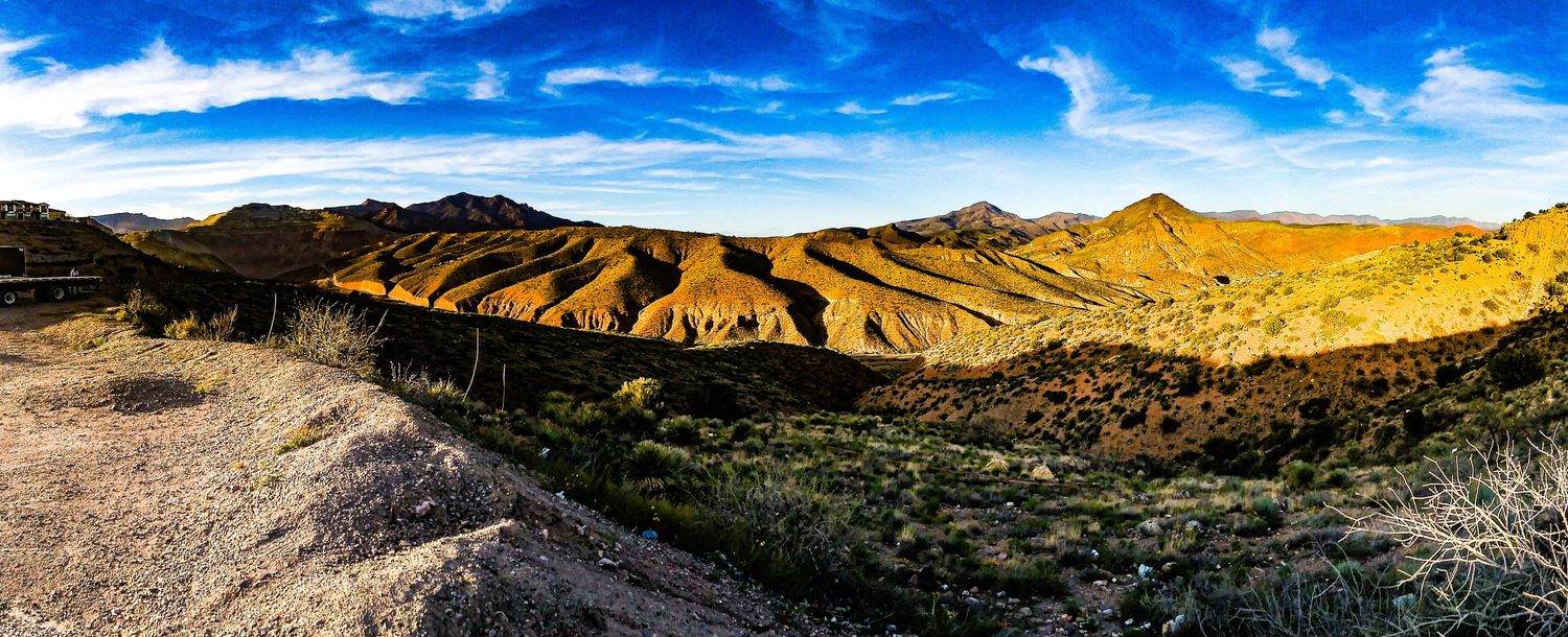 Panoramic desert landscape with rolling golden hills and mountains under bright blue sky.