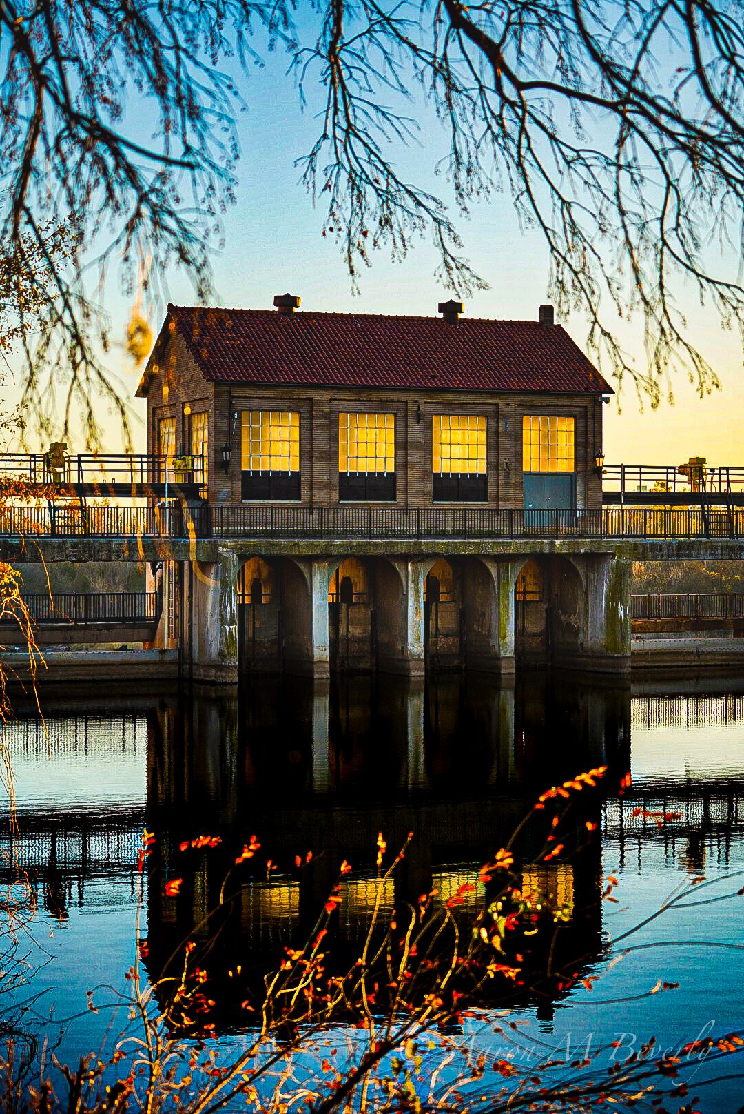 Historic brick pump house with glowing windows reflects perfectly in calm water at sunset.