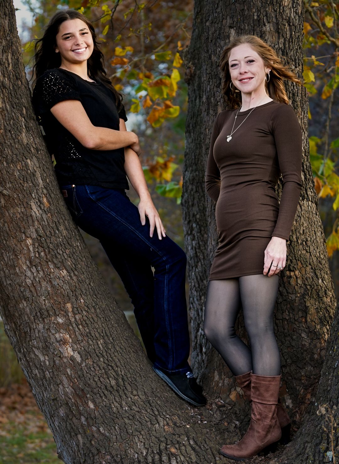 Two women in casual outfits leaning against a tree trunk surrounded by fall foliage.