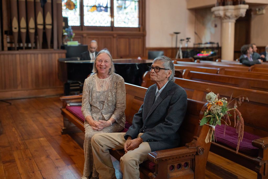 Two people sit together smiling during a wedding on wooden church pews