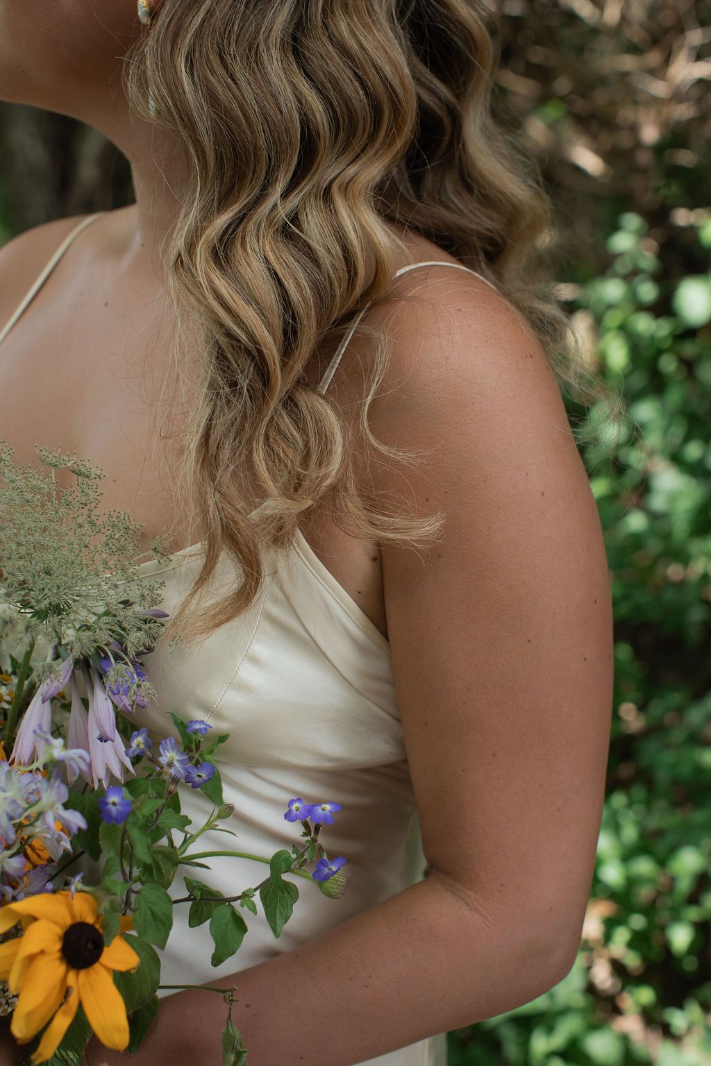 A person in a white dress holds a bouquet with sunflowers and purple wildflowers against a garden background