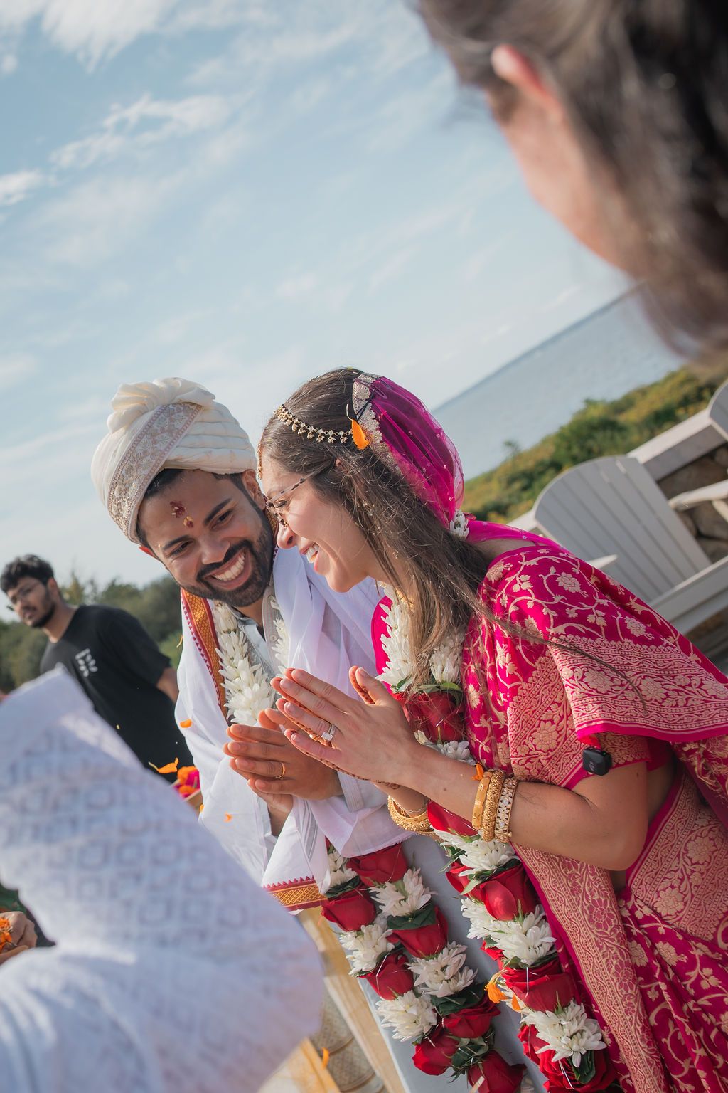 indian bride and groom smile at each other on a sunny day with the ocean behind them