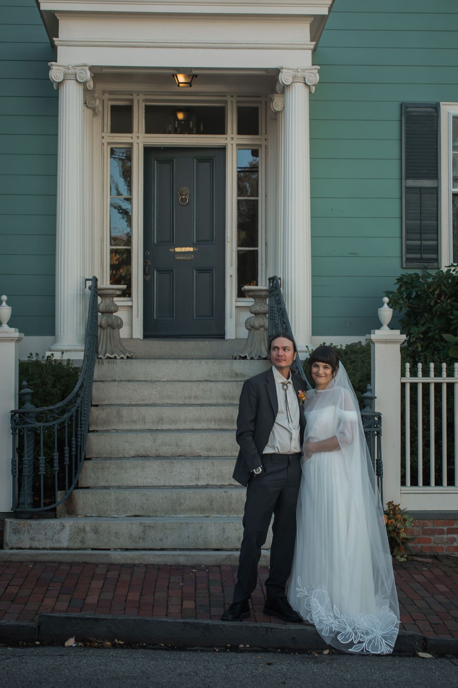 A couple poses in front of a teal-colored historic house with white trim and columns during their wedding day.