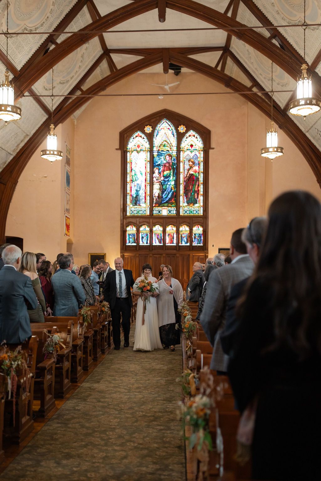 Wedding ceremony in a small church with stained glass windows and wooden pews decorated with floral arrangements.