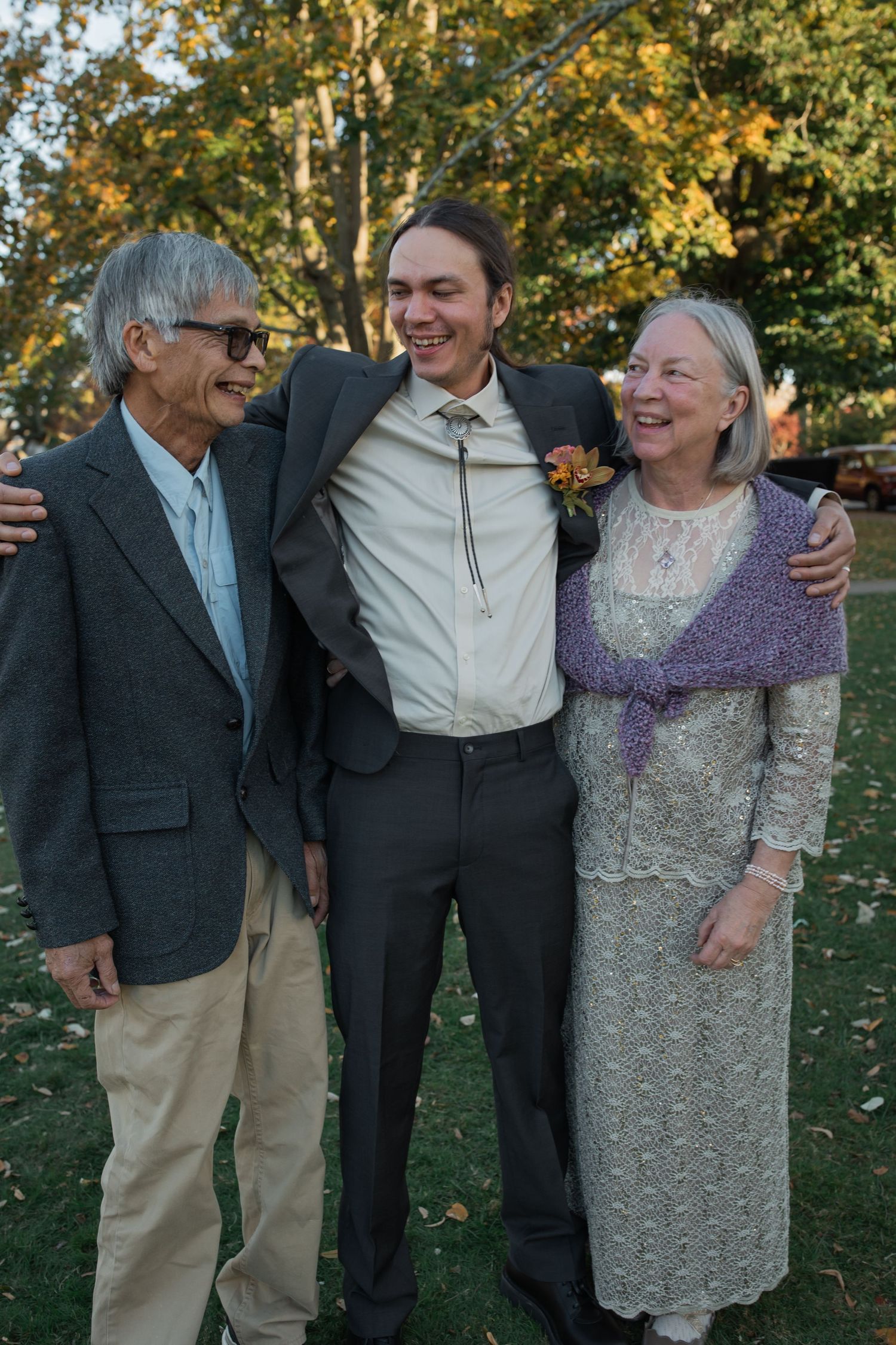 Candid photo showing three people laughing and embracing outdoors during a formal event in autumn.