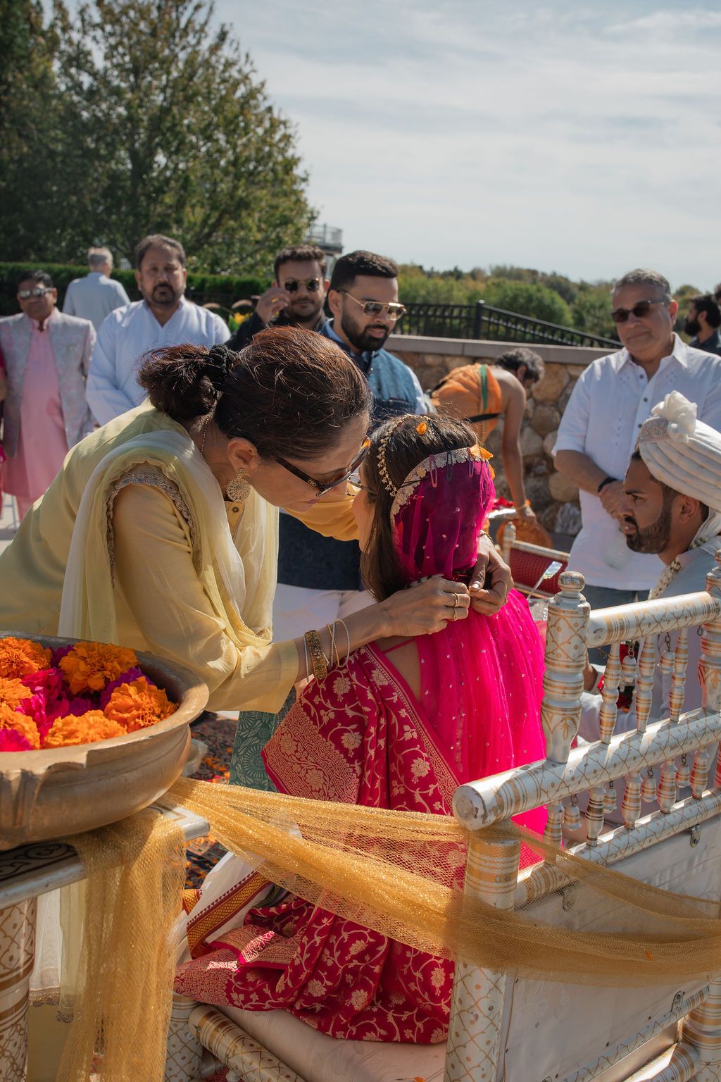 woman in yellow sari attaches necklace to woman in a pink sari sitting in a chair