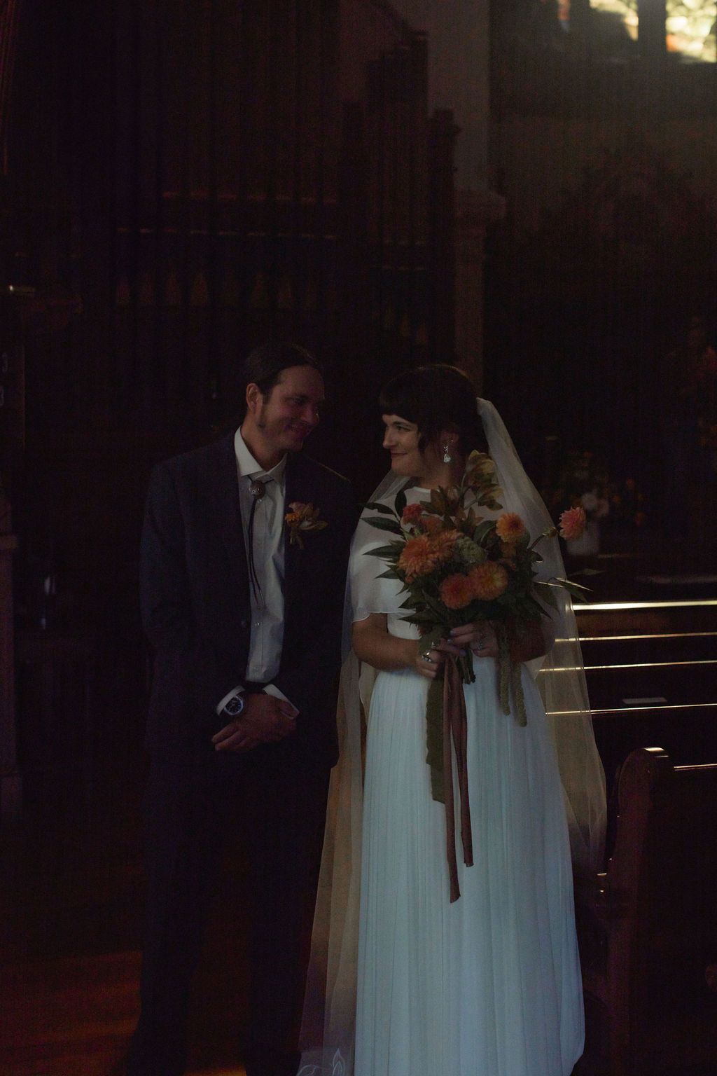 A wedding couple stands together in a dimly lit church interior with stained glass windows and wooden pews.