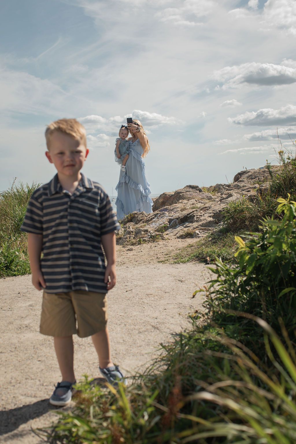 A young child in a striped polo shirt and khaki shorts stands on a rocky coastal path on a sunny day.