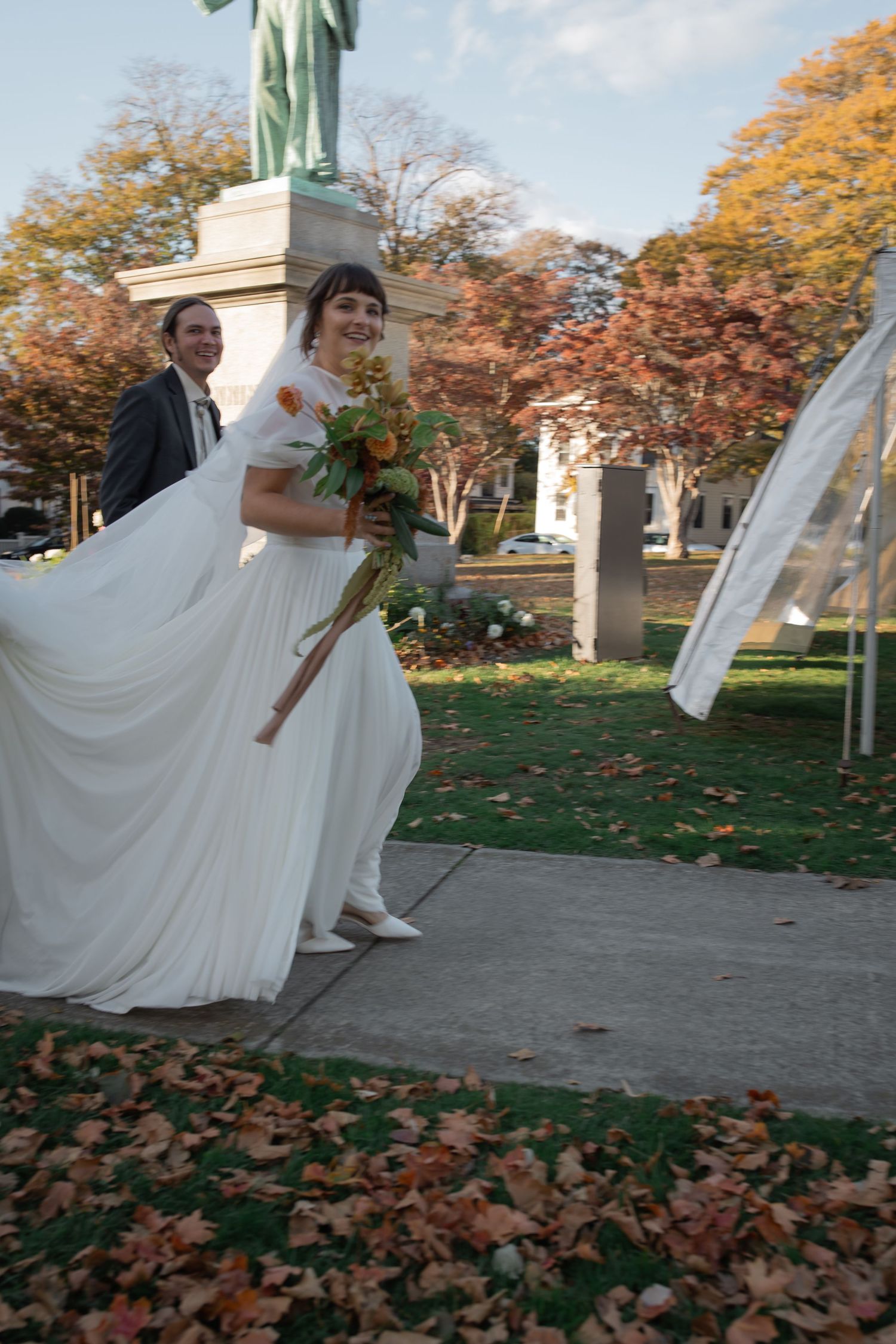 A bride's white dress flows dramatically in the wind as autumn leaves scatter across a garden pathway.
