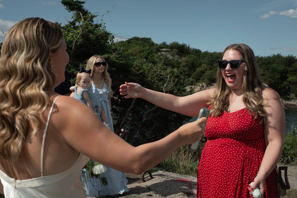 A person in a red polka dot dress greets bride in  a sunny outdoor setting with trees and water in the background.