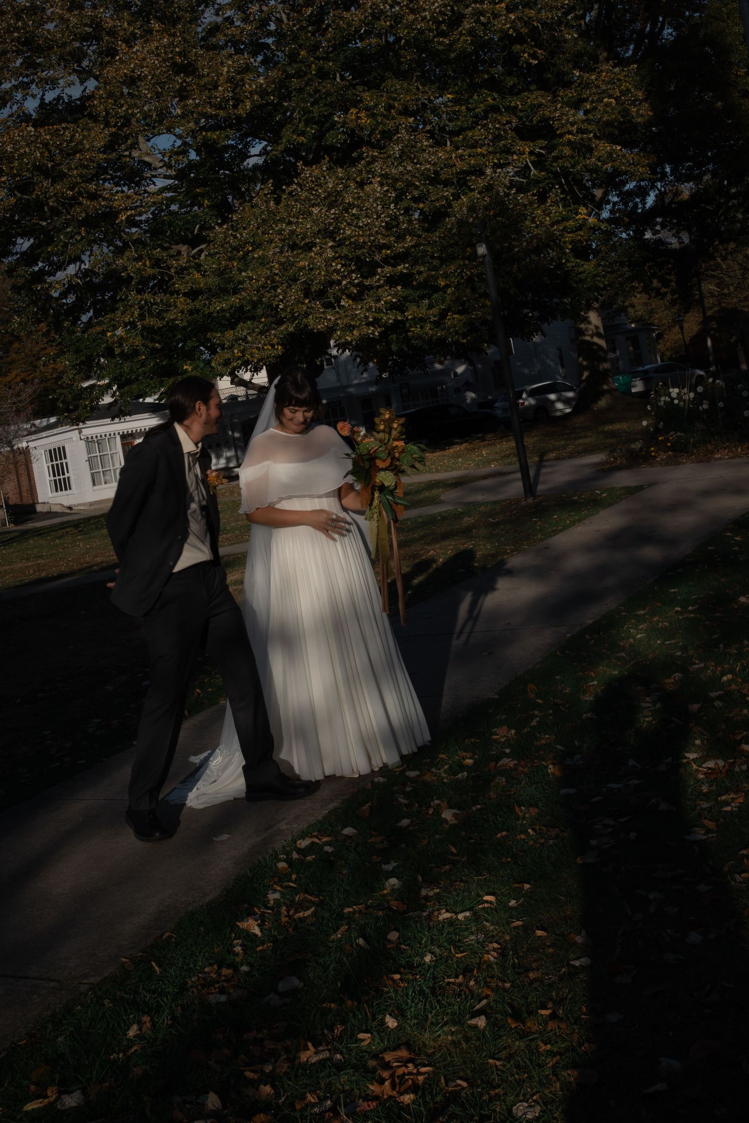 A couple in wedding attire walks together through a park with autumn leaves and shadows on the ground.
