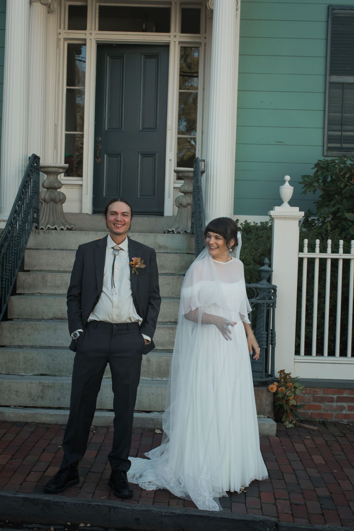 A couple in wedding attire stands on brick steps outside a historic green house with a dark front door.