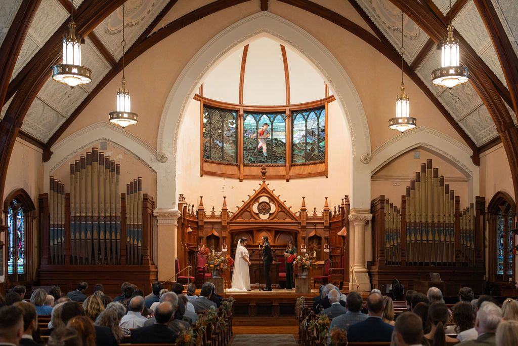 Interior panorama of a church sanctuary with stained glass windows, wooden pews, and hanging pendant lights.