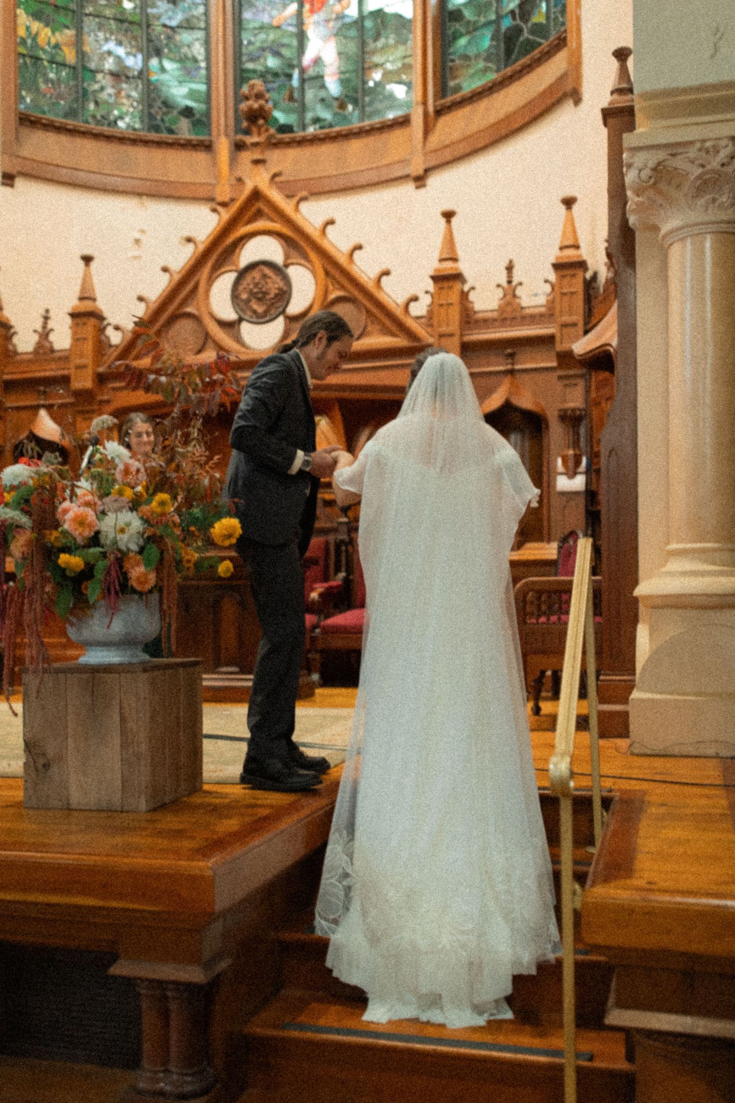 Bride and groom walk up the straits to the alter in a beautiful church chapel with ornate wooden pews and stained glass windows.