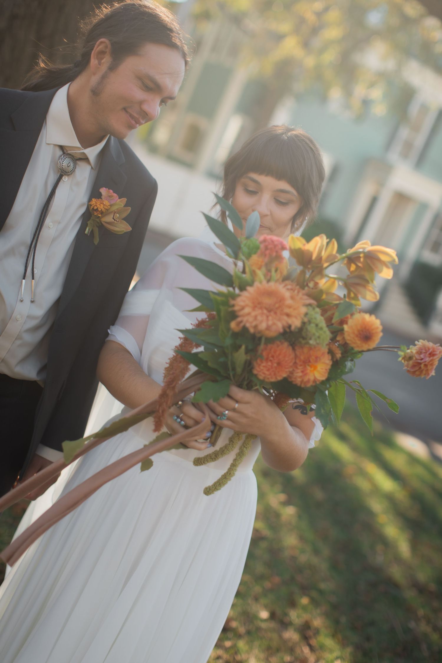 A couple in wedding attire admires a vibrant autumn bouquet with orange dahlias and flowers in warm sunlight.