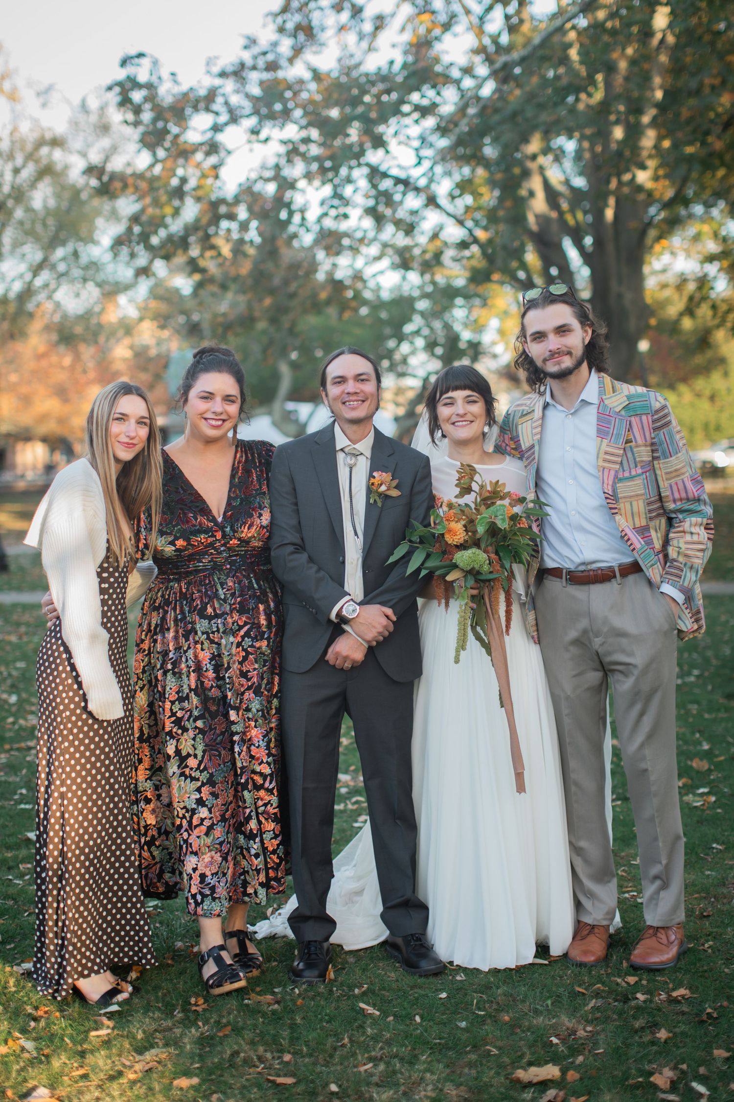 Wedding party stands together outdoors in autumn with mix of casual and formal attire in coordinated earth tones.