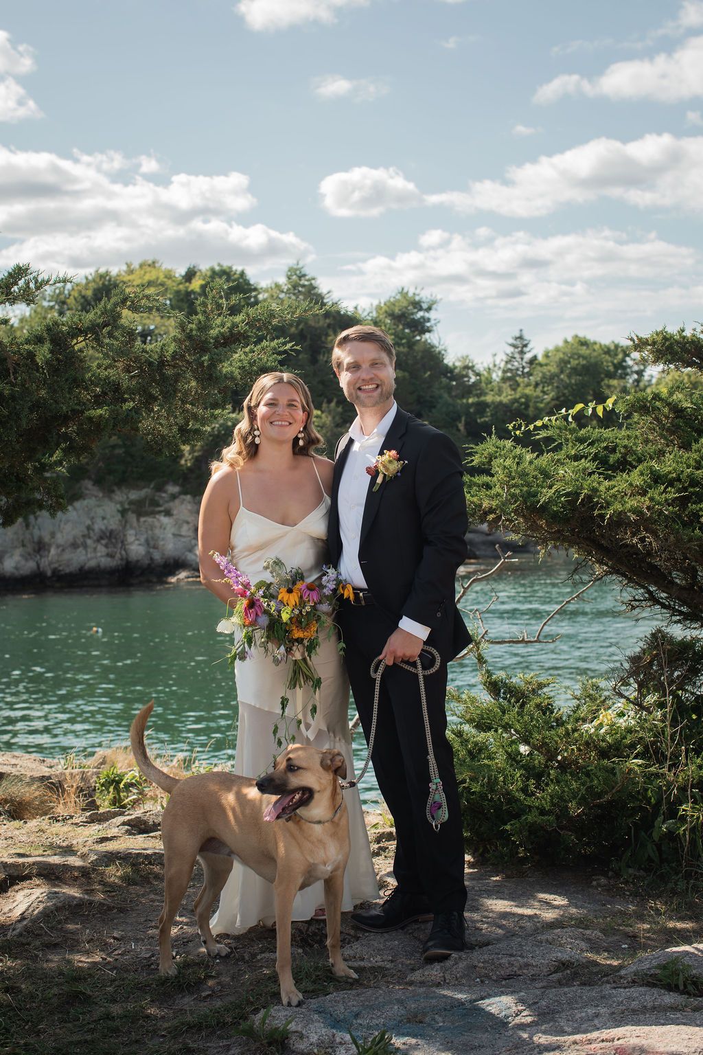 A couple and their dog pose together on a rocky cliff overlooking turquoise waters on a sunny day with white clouds.