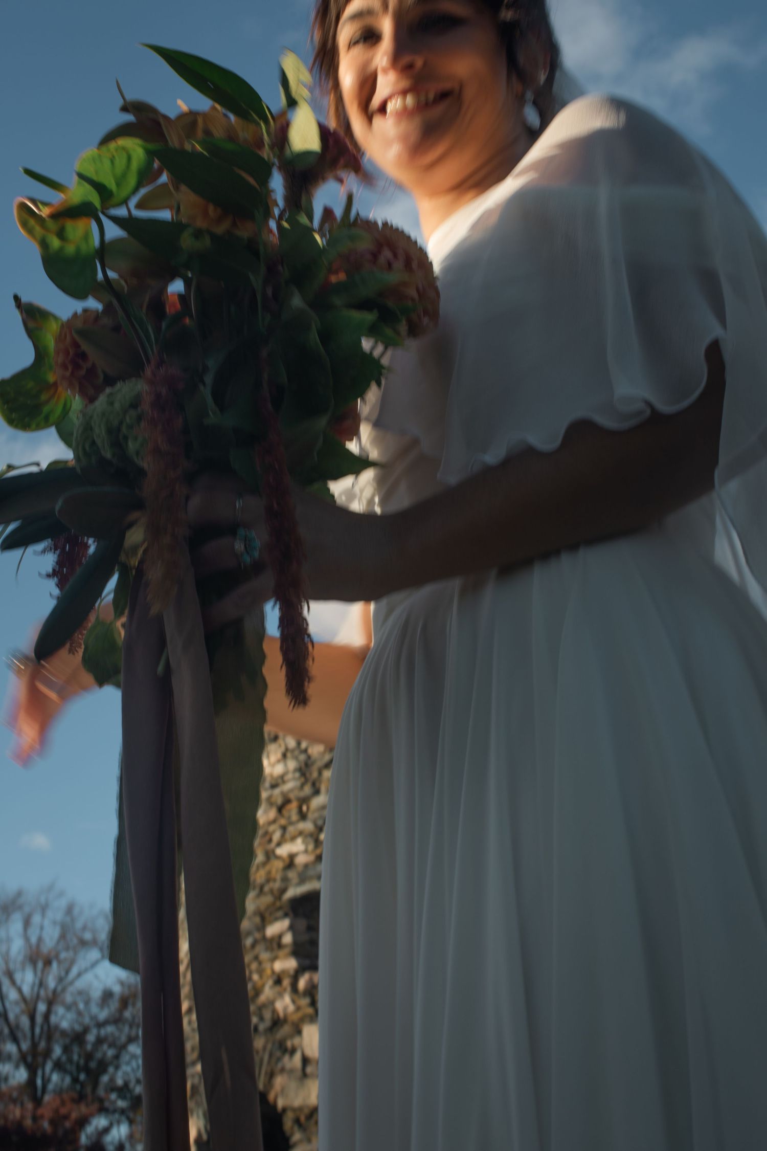 Bride with flowing white dress holds a vibrant floral bouquet against a blue sky backdrop.