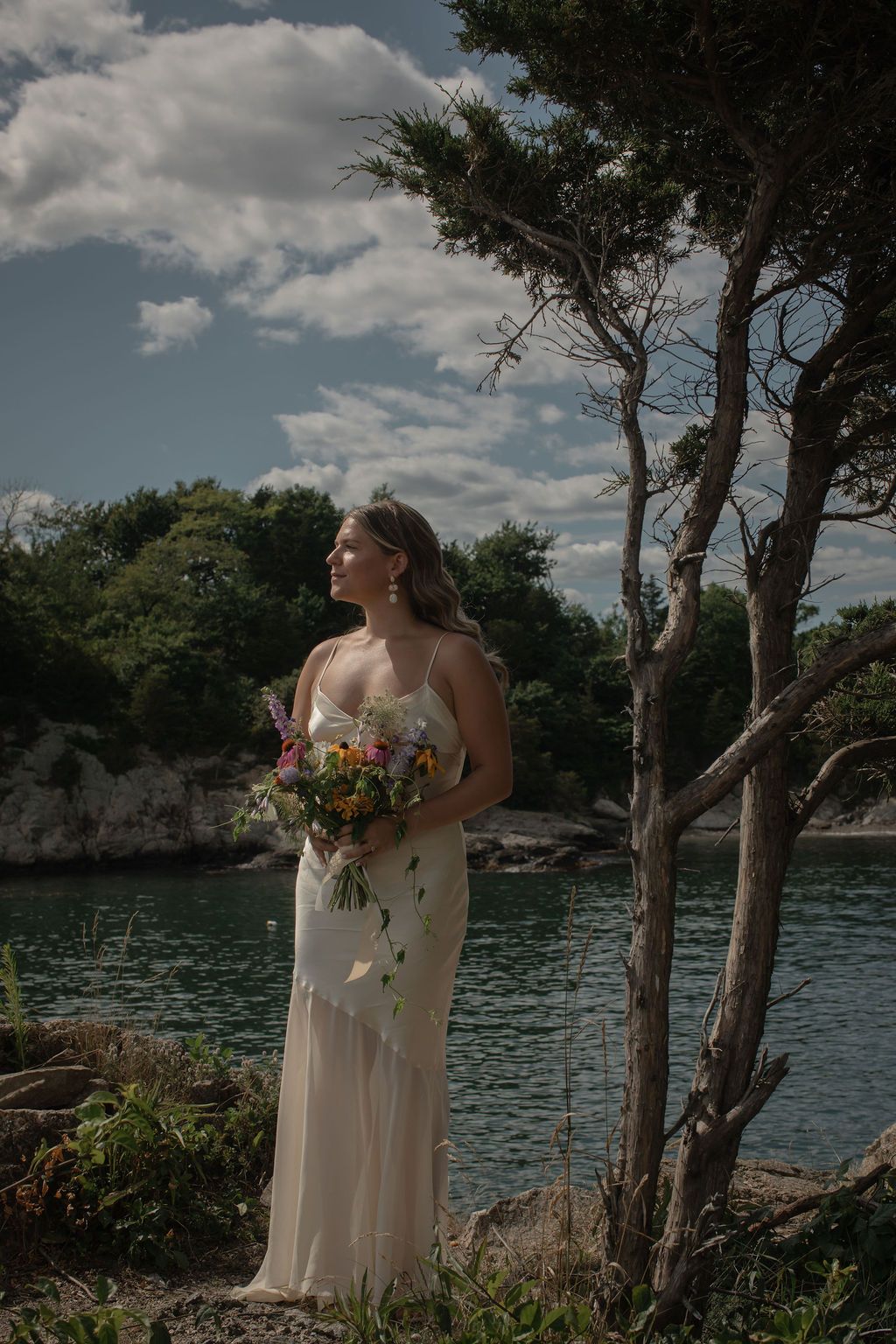 A bride in a flowing cream dress holds a vibrant bouquet while standing on rocky cliffs overlooking a serene lake.