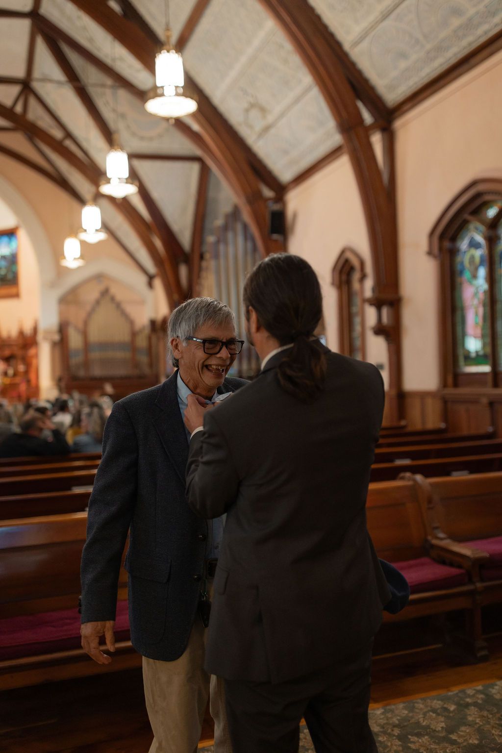 groom pins flowers on his dad's lapel inside a historic church with stained glass windows and wooden arches in Newport, Rhode Island