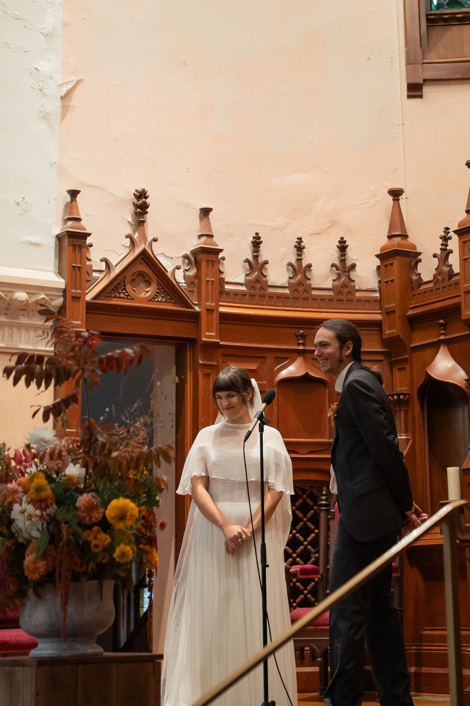 Wedding ceremony taking place in a historic chapel with ornate wooden paneling and floral decorations.