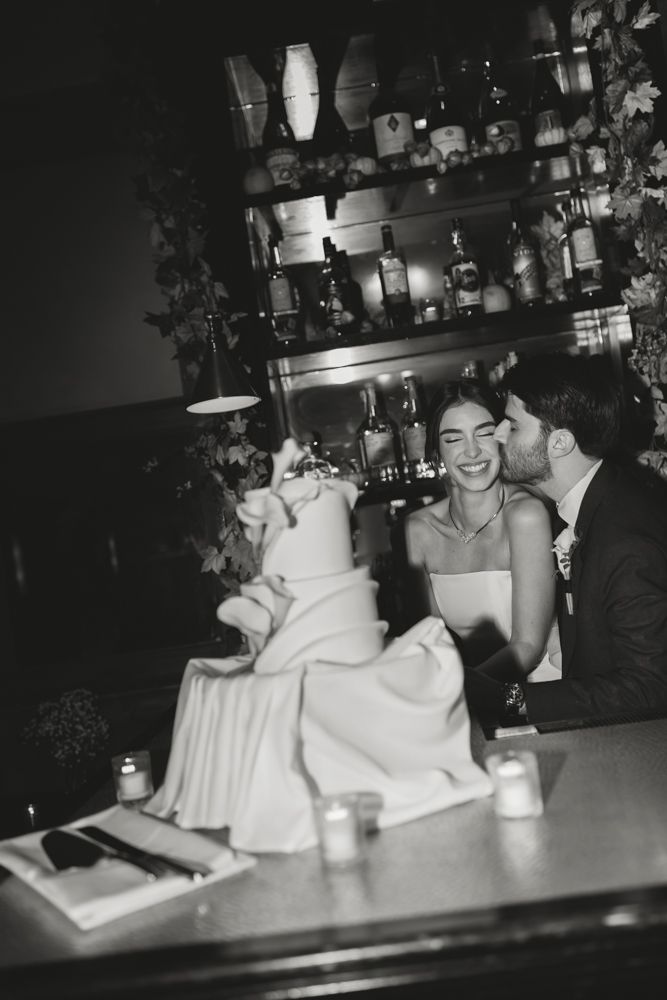 black and white photo of groom kissing bride on the cheek near their wedding cake