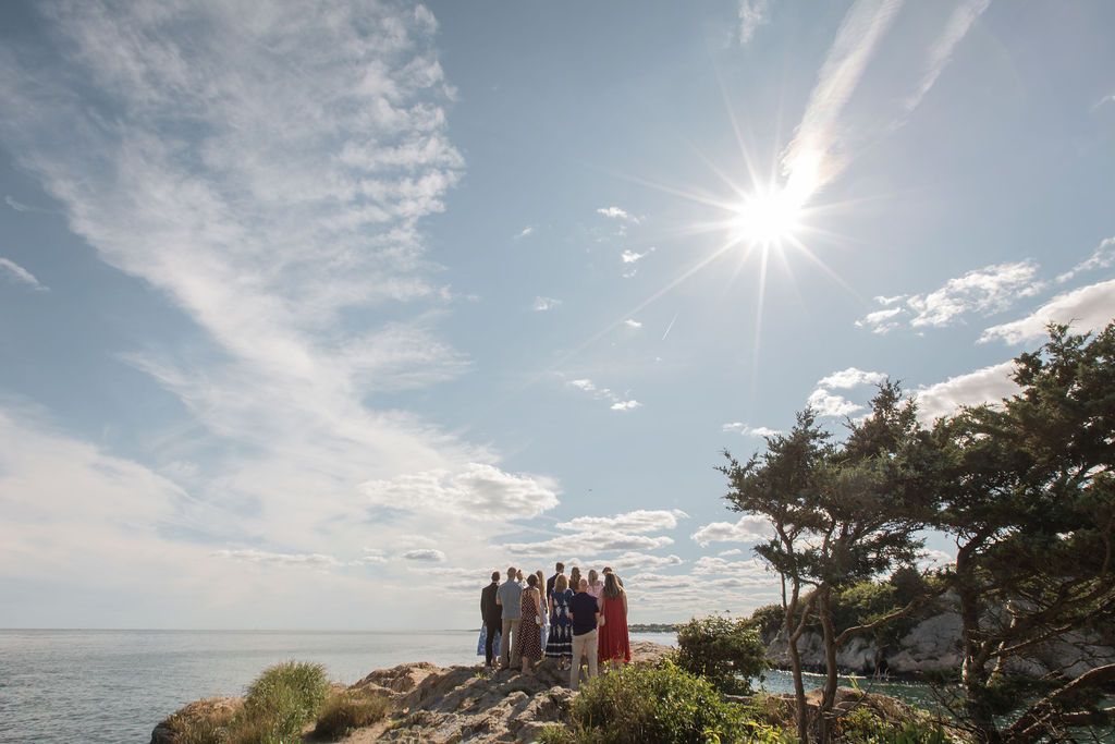 A group of wedding guests enjoying scenic coastal view with bright sunlight streaming through clouds over the ocean and rocky shoreline.