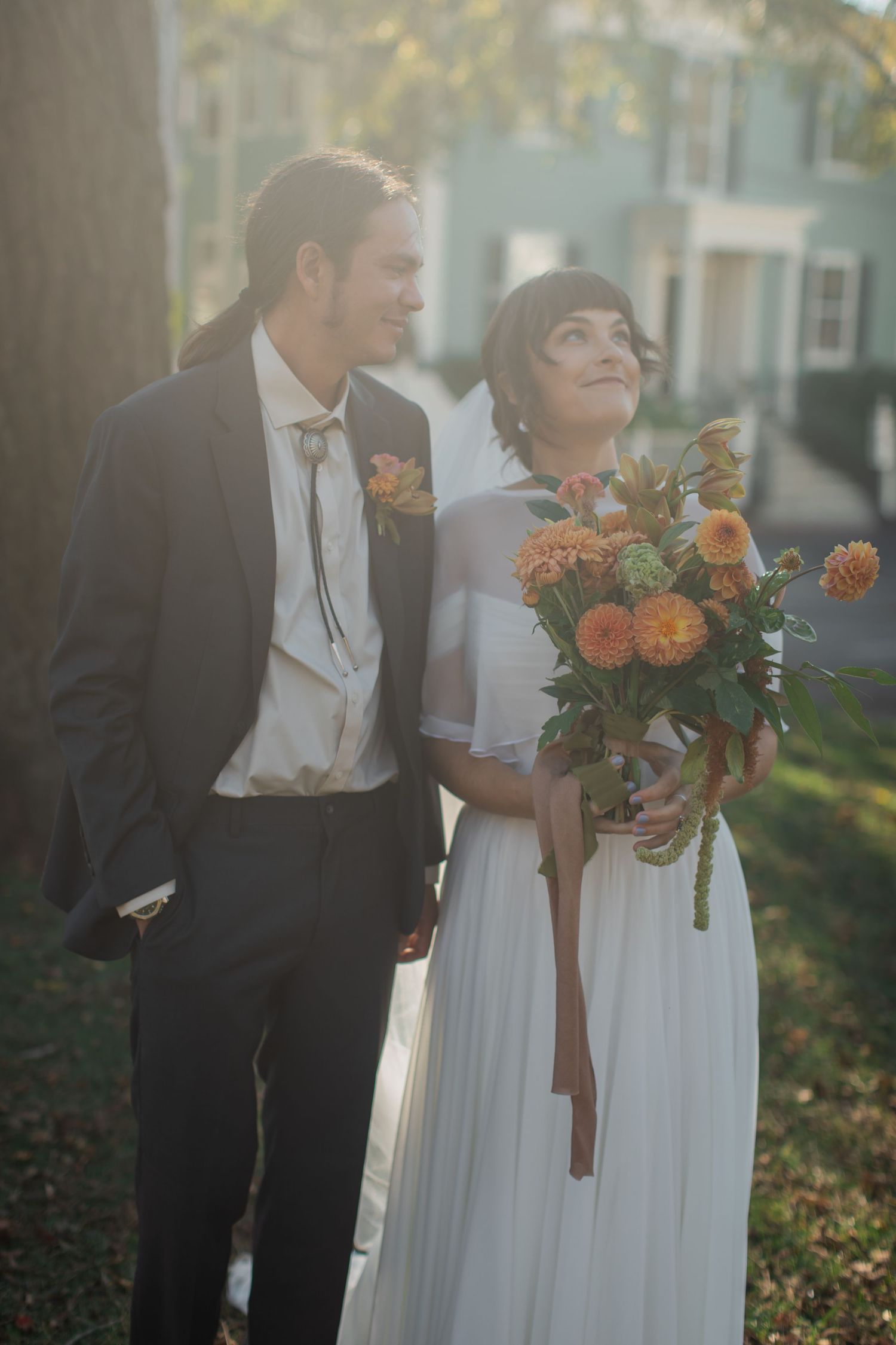 A couple in wedding attire stands together outdoors, with the bride holding a bouquet of orange flowers in golden sunlight.