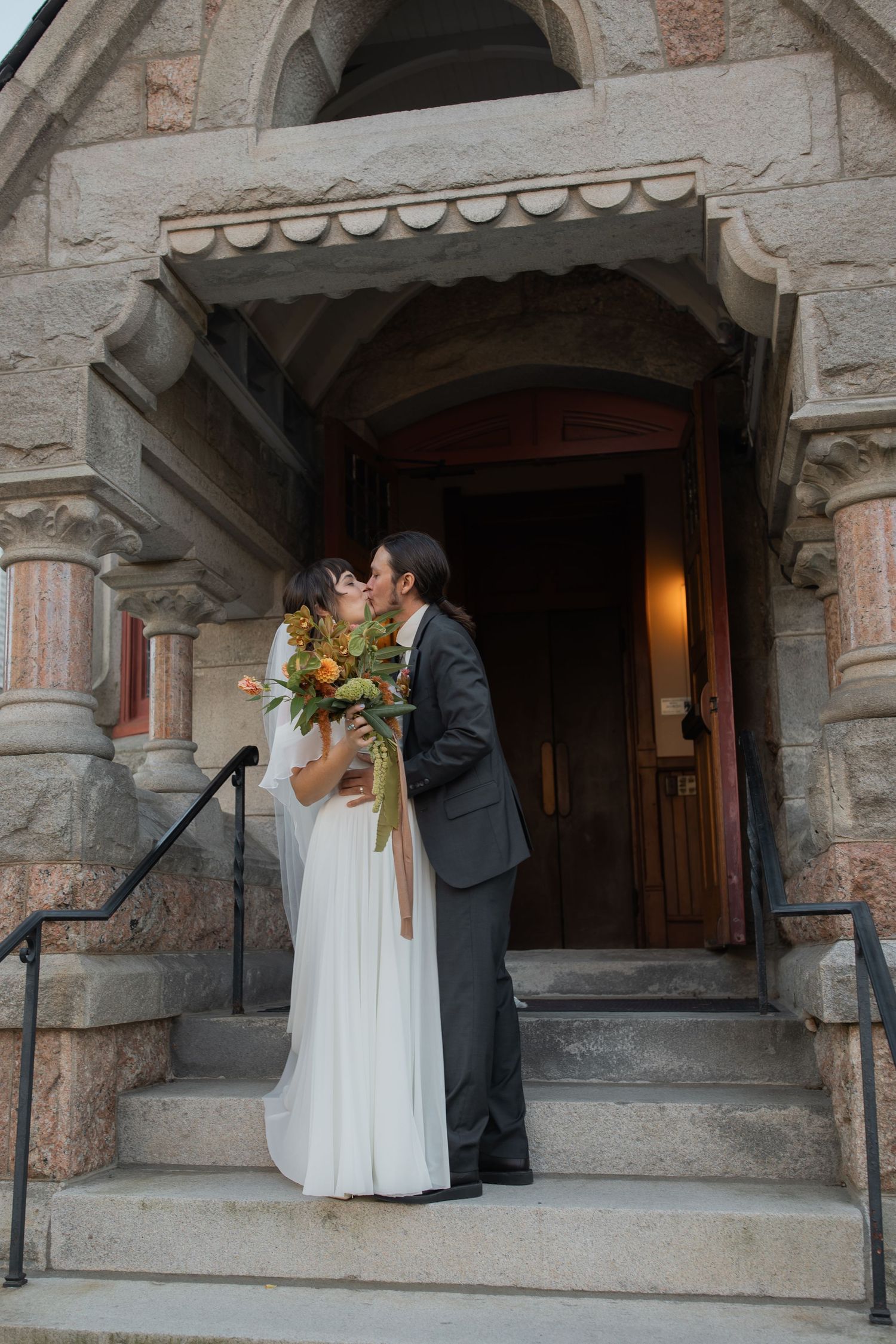 Couple in wedding attire shares intimate moment on stone church steps during sunset ceremony.