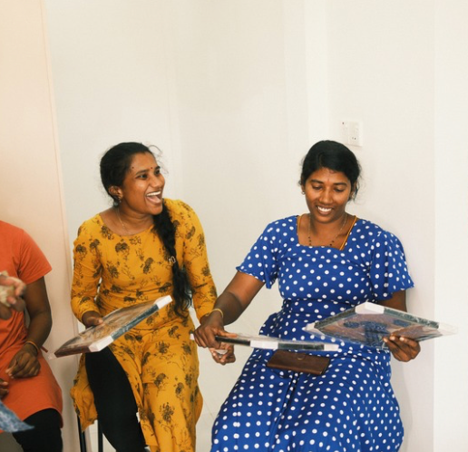 Two women in yellow and polka dot dresses dance and laugh while holding serving trays.